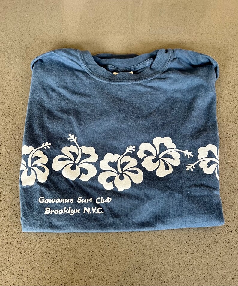 May include: A blue t-shirt with a white hibiscus flower design and the text "Gowanus Surf Club Brooklyn N.Y.C."