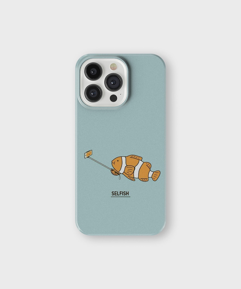 Op de afbeelding: Een lichtblauwe telefoonhoes met een cartoonillustratie van een oranje en witte vis die een selfie maakt met een selfie-stick. De tekst "SELFISH" staat onder de vis.