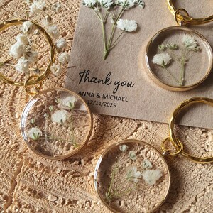 May include: Gold keychains with small, clear, round resin charms containing dried white baby's breath flowers. The keychains are on a brown surface with a thank you card that reads "Thank you Anna & Michael 12/11/2025".