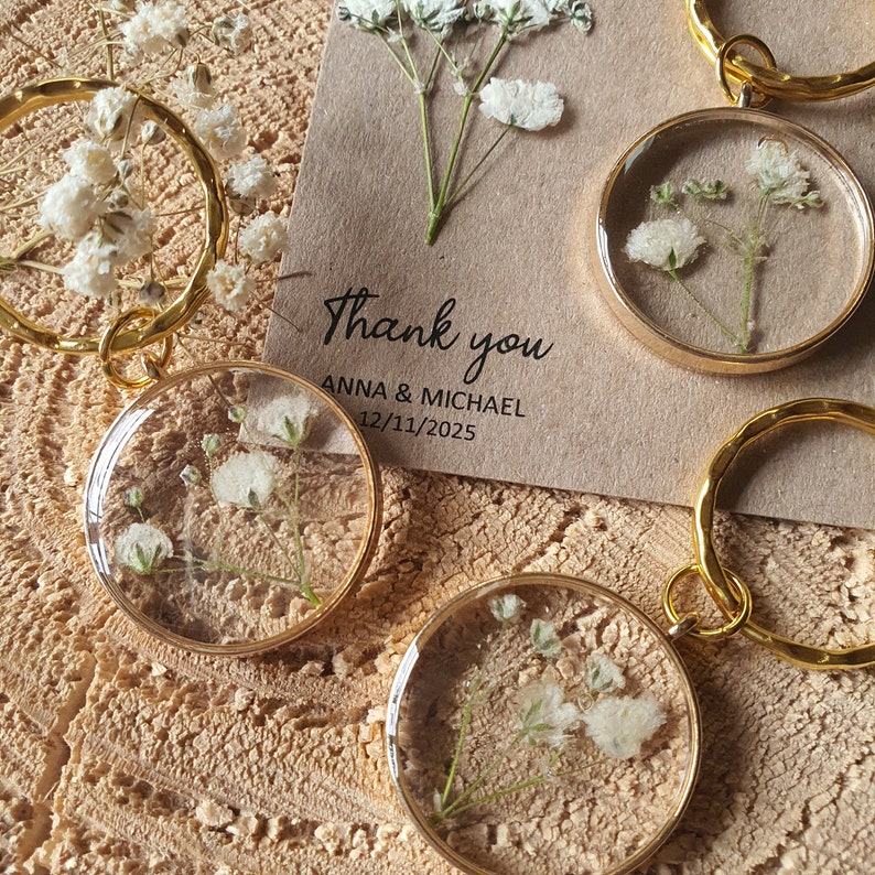 May include: Gold keychains with small, clear, round resin charms containing dried white baby's breath flowers. The keychains are on a brown surface with a thank you card that reads "Thank you Anna & Michael 12/11/2025".