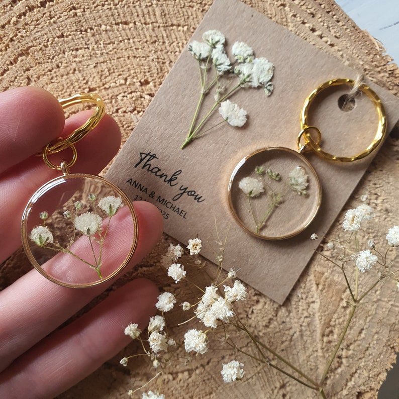 May include: Two gold keychains with clear resin circles containing pressed white baby's breath flowers. The keychains are on a brown wooden surface with a thank you tag that says "Thank you Anna & Michael 12.12.2025".