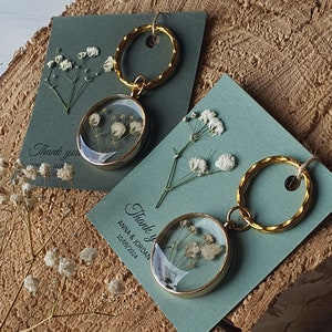 Può includere: Due portachiavi dorati con un ciondolo rotondo di vetro contenente fiori bianchi secchi. I ciondoli sono attaccati a un anello di metallo dorato. I portachiavi sono esposti su una carta blu con il testo "Grazie Anna & Jordan 10/09/2024".