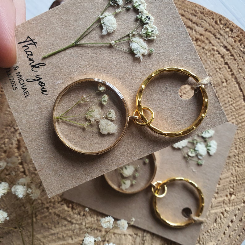 May include: Gold keychains with a small round charm featuring pressed white flowers inside clear resin. The charms are attached to a gold ring. The keychains are displayed on a brown paper card with the text "Thank you & Michael July 2025".