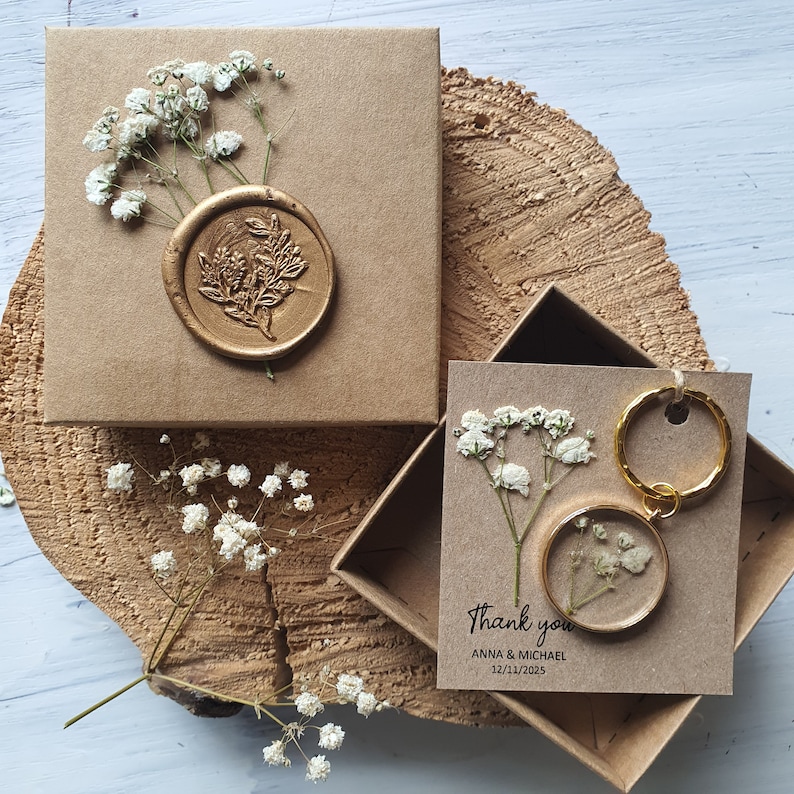 May include: Two small kraft paper boxes with a gold wax seal on one box and a small dried flower arrangement inside a clear resin keychain on the other. The boxes are on a wooden surface. The text on the box reads "Thank you Anna & Michael 12/11/2025".