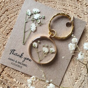 May include: A gold key chain with a round, clear resin charm containing dried white flowers. The keychain is on a brown card with the text "Thank you Anna & Michael 12/11/2025".