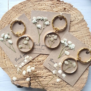 May include: Set of three gold keychains with small glass circles containing pressed white flowers. Each keychain has a brown card attached with the text "Thank you" and the names "Anna & Michael" and the date "12/11/2025".