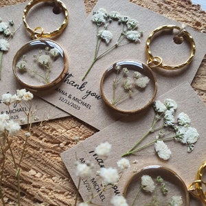 May include: Gold keychains with small, clear, round resin charms containing dried white baby's breath flowers. The keychains are attached to small cards with the text "Thank you" and the names "Anna & Michael" and the date "12/11/2025".