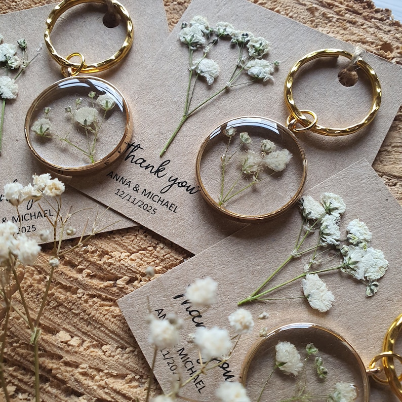 May include: Gold keychains with small, clear, round resin charms containing dried white baby's breath flowers. The keychains are attached to small cards with the text "Thank you" and the names "Anna & Michael" and the date "12/11/2025".