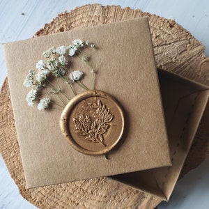 May include: A brown cardboard box with a gold wax seal. The seal has a floral design. The box is decorated with white flowers.