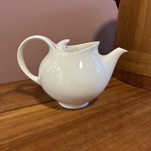 May include: A white ceramic teapot with a curved handle and spout. The teapot has a rounded body and a small, decorative finial on the lid. It sits on a wooden surface, with a wooden bucket in the background.