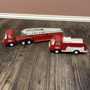May include: Three vintage Tonka fire trucks. The trucks are red with white accents and black wheels. One truck has a ladder, and another has a white hose reel. The trucks are on a wooden surface.
