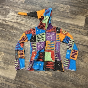 May include: A patchwork hoodie with a pointed hood and bell sleeves. The jacket features a variety of colorful squares in shades of blue, orange, purple, and green. The jacket has a zipper closure and is made of a soft, textured fabric.