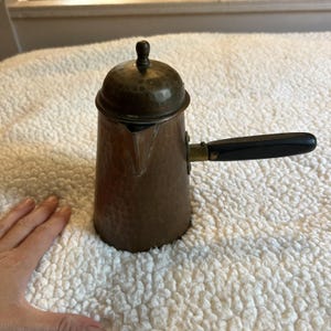 May include: A copper-colored coffee pot with a hammered texture, a lid, and a black handle. The pot has a spout and a small knob on the lid. The handle is attached with a brass fitting. The pot is likely vintage and is designed for stovetop use.
