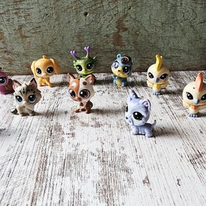 May include: A collection of colorful, small plastic animal figurines, including cats, dogs, and birds, with large eyes. The toys are arranged on a distressed white wooden surface, with a weathered green backdrop.