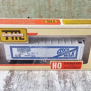 Peut inclure: Une maquette de wagon de train à l'échelle HO vintage dans sa boîte d'origine. La boîte est crème et orange avec les mots "Train-Miniature" et "HO Railroad Kits and Accessories". Le wagon est blanc avec des lettres bleues qui disent "American" et "Cisco Milk".