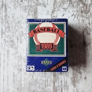 Puede incluir: Una taza de cerámica blanca con un asa negra y el texto "But First, Coffee" en negrita negra. La taza se encuentra sobre un fondo borroso, sugiriendo un ambiente acogedor.