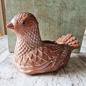 May include: A terracotta bird-shaped planter with detailed carved feather and scale patterns. The bird has a detailed head with a beak and an eye. The planter is a warm brown colour with white accents, suitable for small plants or decorative use.