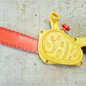 May include: A vintage toy chainsaw in bright yellow and red. The word "SAW" is prominently displayed on the yellow body. The red blade has a scalloped edge, and the toy features a red handle and a small red knob.