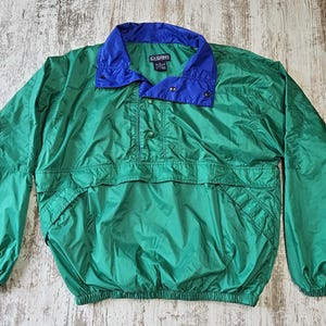 May include: A green and blue anorak jacket. The jacket has a green body with a blue collar and accents. It features a half-zip closure, a front pocket, and elastic cuffs. The jacket is made of a lightweight, possibly nylon, material.