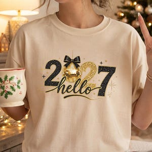 Puede incluir: Una camiseta beige de cuello redondo con un gráfico que dice hello 2027, donde una bola de discoteca dorada reemplaza el cero. El diseño incluye detalles en negro y purpurina dorada, estrellas y un lazo negro. Se sostiene una taza blanca con un patrón de acebo.