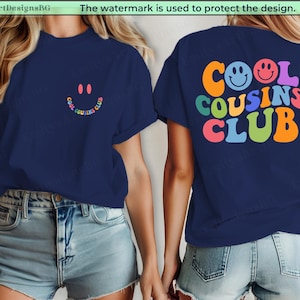 May include: Navy blue t-shirt featuring a smiley face and the text "COOL COUSINS CLUB" in colourful, retro-style lettering. The design is on the front and back of the shirt. The shirt is worn with denim shorts.