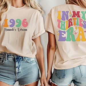 May include: Beige t-shirt featuring the year "1996" in orange, purple, and green, and the text "Hannah's Version." The back of the shirt displays "In My Thirties Era" in colourful, retro-style lettering. The shirt is worn with denim shorts.