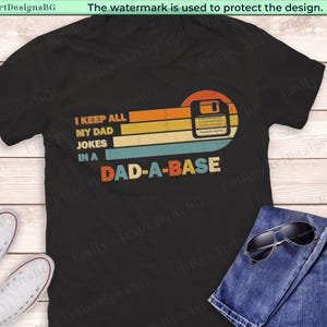 Könnte beinhalten: Schwarzes T-Shirt mit Retro-Design, das eine Diskette und den Text "I KEEP ALL MY DAD JOKES IN A DAD-A-BASE" zeigt. Das Shirt wird mit einer blauen Jeans und Sonnenbrille kombiniert.