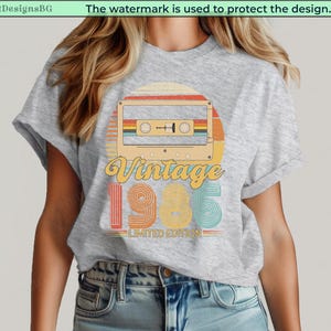May include: A light gray t-shirt featuring a retro cassette tape design with the words "Vintage 1985 Limited Edition." The design incorporates a sunset color scheme with orange, yellow, and teal accents, evoking a nostalgic feel.