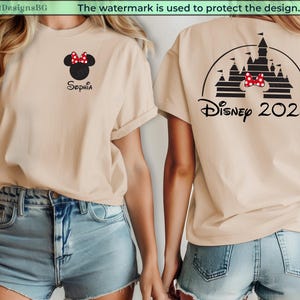 Puede incluir: Camiseta beige con dise&ntilde;o de Minnie Mouse, lazo rojo y el nombre "Sophia". La parte trasera muestra un castillo y "Disney 2026". La camiseta se combina con shorts de mezclilla. La imagen muestra un atuendo casual.