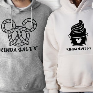May include: Two gray and white hoodies with a black graphic design. One hoodie has a pretzel shaped like Mickey Mouse ears with the text "Kinda Salty". The other hoodie has a black ice cream cone with a Mickey Mouse head on the bottom and the text "Kinda Sweet".