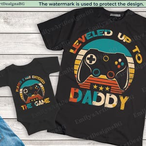 Puede incluir: Una camiseta negra con un gráfico retro de un mando de videojuego y el texto "LEVELED UP TO DADDY". Un body negro con un gráfico retro de un mando de videojuego y el texto "PLAYER 2 HAS ENTERED THE GAME".