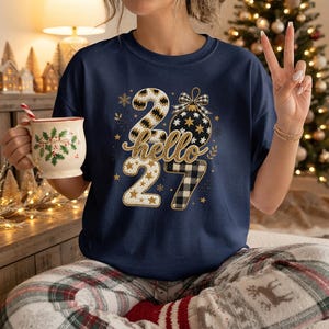 Puede incluir: Una camiseta azul marino con un diseño gráfico que incluye el texto hello 2027. Los números están decorados con patrones de cuadros negros y blancos, estrellas doradas y un adorno navideño colgante con un lazo y copos.
