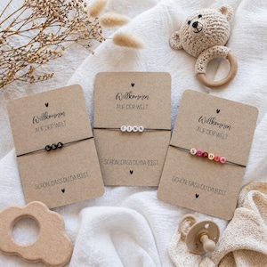 May include: Three personalized bracelets with names spelled out in colorful beads are displayed on kraft paper cards. Each card has the text "Willkommen auf der Welt" and "Sch&ouml;n, dass du da bist". A wooden toy, a pacifier, and a crocheted bear rattle are also visible.