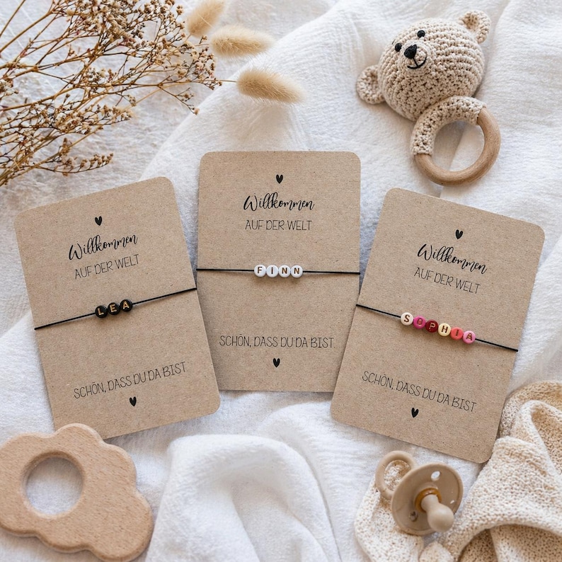 May include: Three personalized bracelets with names spelled out in colorful beads are displayed on kraft paper cards. Each card has the text "Willkommen auf der Welt" and "Sch&ouml;n, dass du da bist". A wooden toy, a pacifier, and a crocheted bear rattle are also visible.