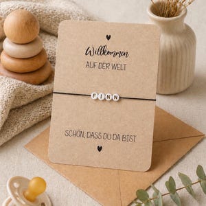 May include: A greeting card with the name "Finn" on a bracelet. The card reads "Willkommen auf der Welt" and "Sch&ouml;n, dass du da bist". A wooden stacking toy, a vase, and a pacifier are also visible.
