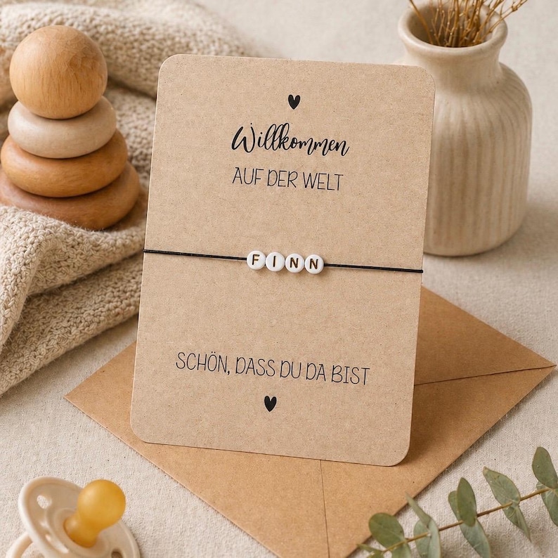 May include: A greeting card with the name "Finn" on a bracelet. The card reads "Willkommen auf der Welt" and "Sch&ouml;n, dass du da bist". A wooden stacking toy, a vase, and a pacifier are also visible.
