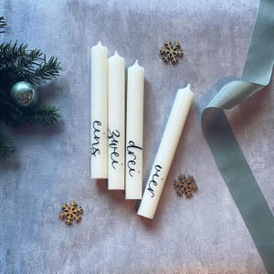 Könnte beinhalten: Vier weiße Kerzen mit schwarzer Kalligraphie auf jeder. Die Kerzen sind mit "eins", "zwei", "drei" und "vier" beschriftet.