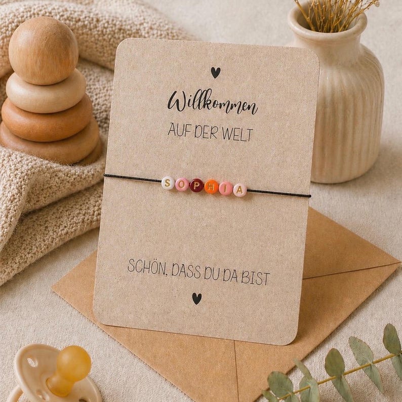 May include: A personalized bracelet with the name "SOPHIA" spelled out in colorful beads. The bracelet is displayed on a card with German text and a small heart symbol. A wooden stacking toy and a small vase are also in the image.