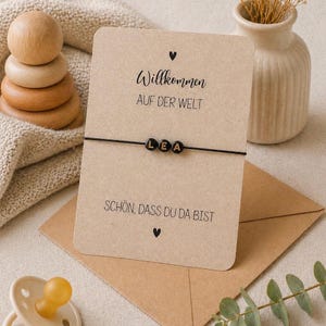 May include: A black beaded bracelet spelling "LEA" on a black string, displayed on a card with German text. The card is on a brown envelope, with a small vase and wooden toy in the background. The text on the card reads "Willkommen auf der Welt" and "Sch&ouml;n, dass du da bist".