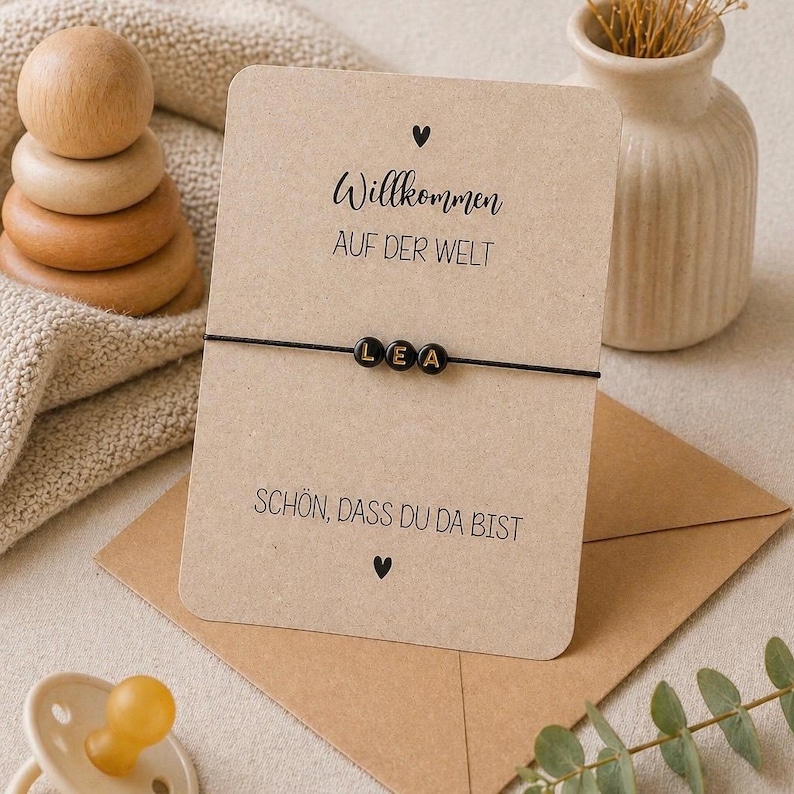 May include: A black beaded bracelet spelling "LEA" on a black string, displayed on a card with German text. The card is on a brown envelope, with a small vase and wooden toy in the background. The text on the card reads "Willkommen auf der Welt" and "Sch&ouml;n, dass du da bist".