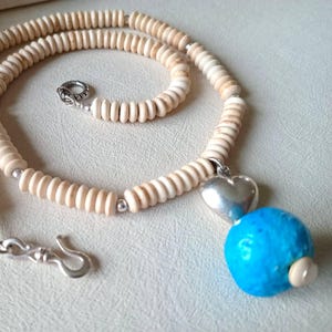 Puede incluir: Un collar de cuentas color crema con un colgante de corazón plateado y una cuenta azul turquesa. El collar tiene un cierre plateado y un cierre circular. Las cuentas son pequeñas y cilíndricas.