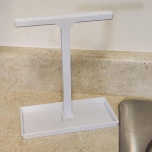 May include: White plastic countertop dish rack with a single bar for drying dishes. The rack has a rectangular base and a vertical bar with a horizontal bar at the top.