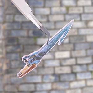 May include: A blue and white sword sticker with a dragon head design. The sticker is being held by tweezers.