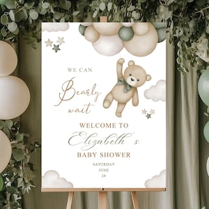 May include: A baby shower sign on a wooden easel, decorated with balloons and greenery. The sign reads "We can bearly wait welcome to Elizabeth's baby shower" with a teddy bear illustration. The balloons are white and green.