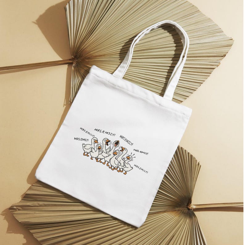May include: White canvas tote bag featuring a cartoon illustration of a flock of geese with the text "Mrs. Emily!". The geese are depicted in a line, with some of them honking. The bag has white handles and is set against a neutral background.