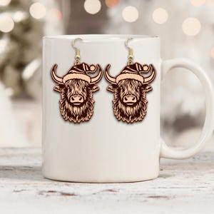 May include: A pair of wooden earrings featuring a Highland cow design wearing a Santa hat. The earrings have gold-colored fish hook ear wires. The cow design is a light brown color with detailed fur and horns, perfect for the holiday season.