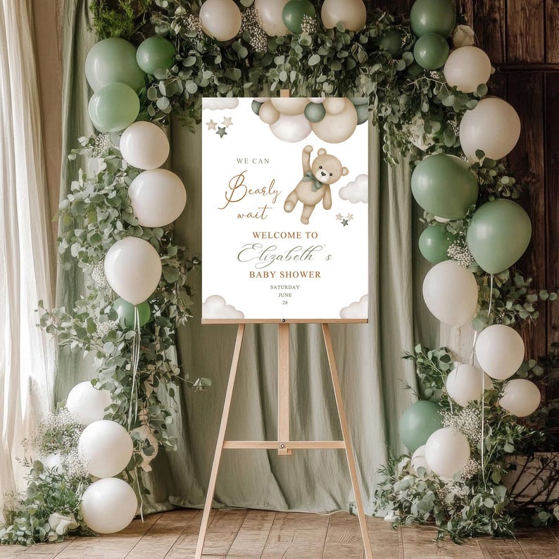 May include: A baby shower sign on a wooden easel, decorated with balloons and greenery. The sign reads "We can bearly wait welcome to Elizabeth's baby shower" with a teddy bear illustration. The balloons are white and green.