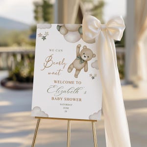 May include: A baby shower welcome sign on a gold easel. The sign features a teddy bear graphic and the text "We can bearly wait welcome to Elizabeth's baby shower". A large cream-coloured bow and fabric drape the side.