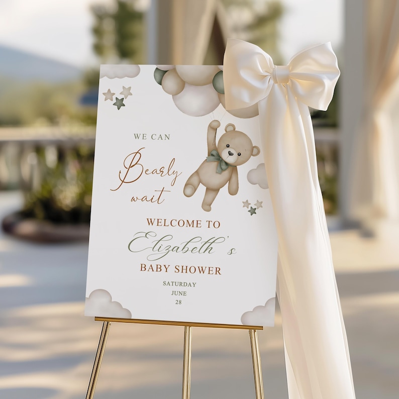 May include: A baby shower welcome sign on a gold easel. The sign features a teddy bear graphic and the text "We can bearly wait welcome to Elizabeth's baby shower". A large cream-coloured bow and fabric drape the side.
