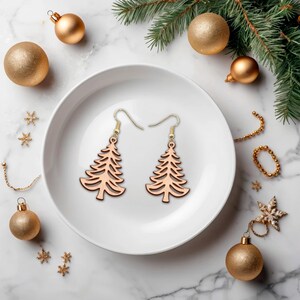 May include: Two wooden Christmas tree earrings with gold-colored hooks rest on a white plate. The earrings are surrounded by gold ornaments, snowflakes, and a sprig of evergreen. The earrings are a festive holiday accessory.